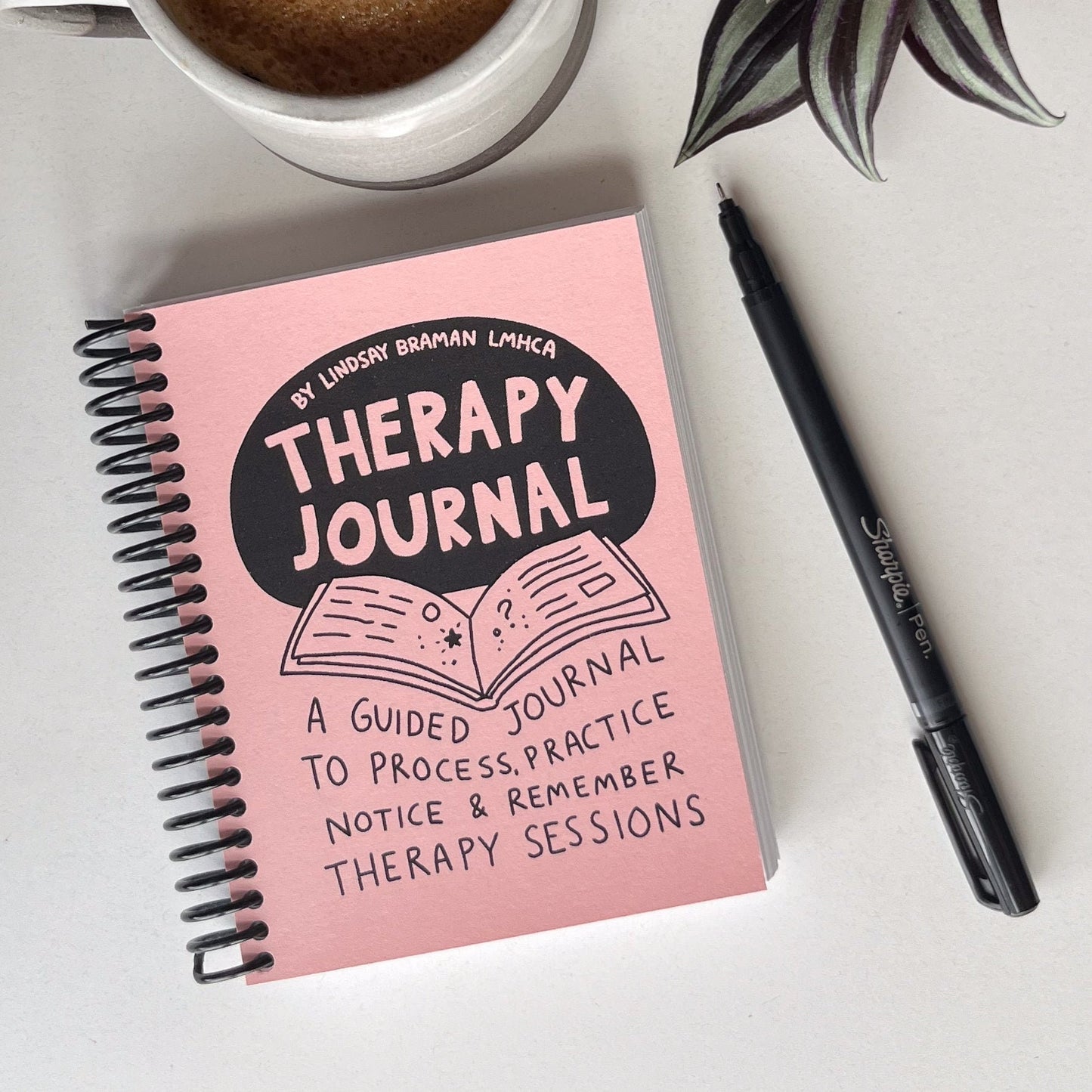 Image of TherapyJournal by Lindsay Braman LMHCA. The journal is palm-sized and spiral bound with a light pink cover.